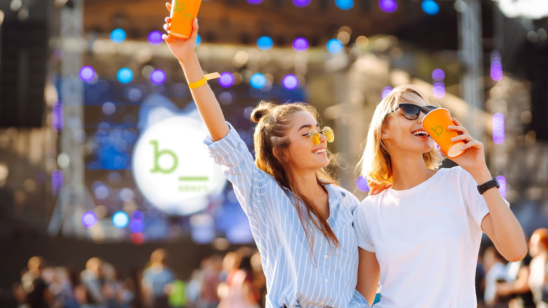 Two young women are holding up orange cups and smiling happily at an outdoor event with a blurred background and a blurred b smart logo.