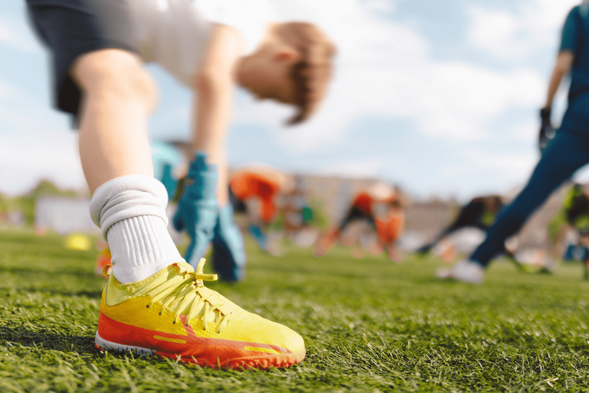 A group of people is stretching on a soccer field with a child in yellow shoes standing in the foreground.