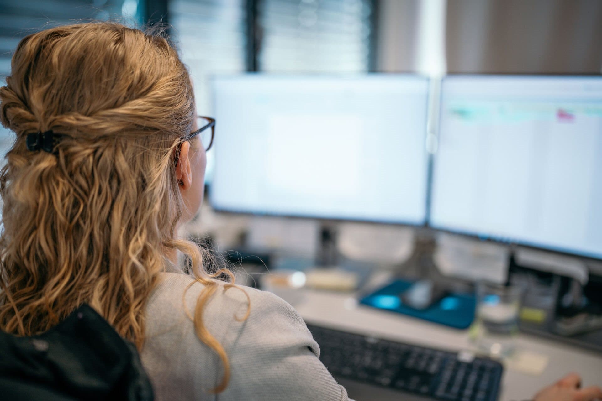 A woman with blonde hair is sitting in an office at a desk working on two computer screens.