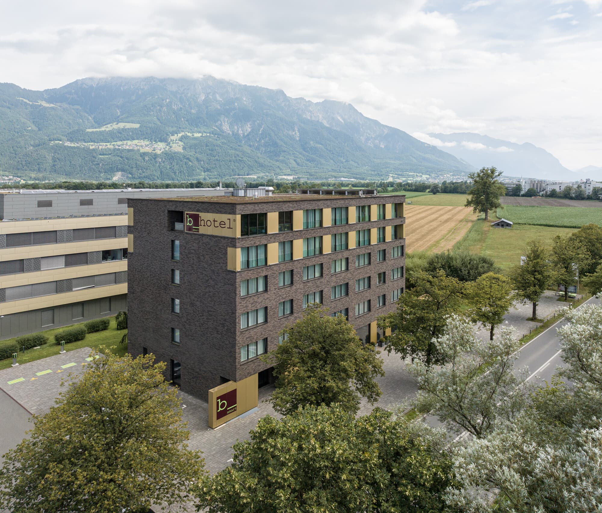A modern multistory hotel building with the sign b smart hotel Bendern stands in a green landscape overlooking forested mountains.