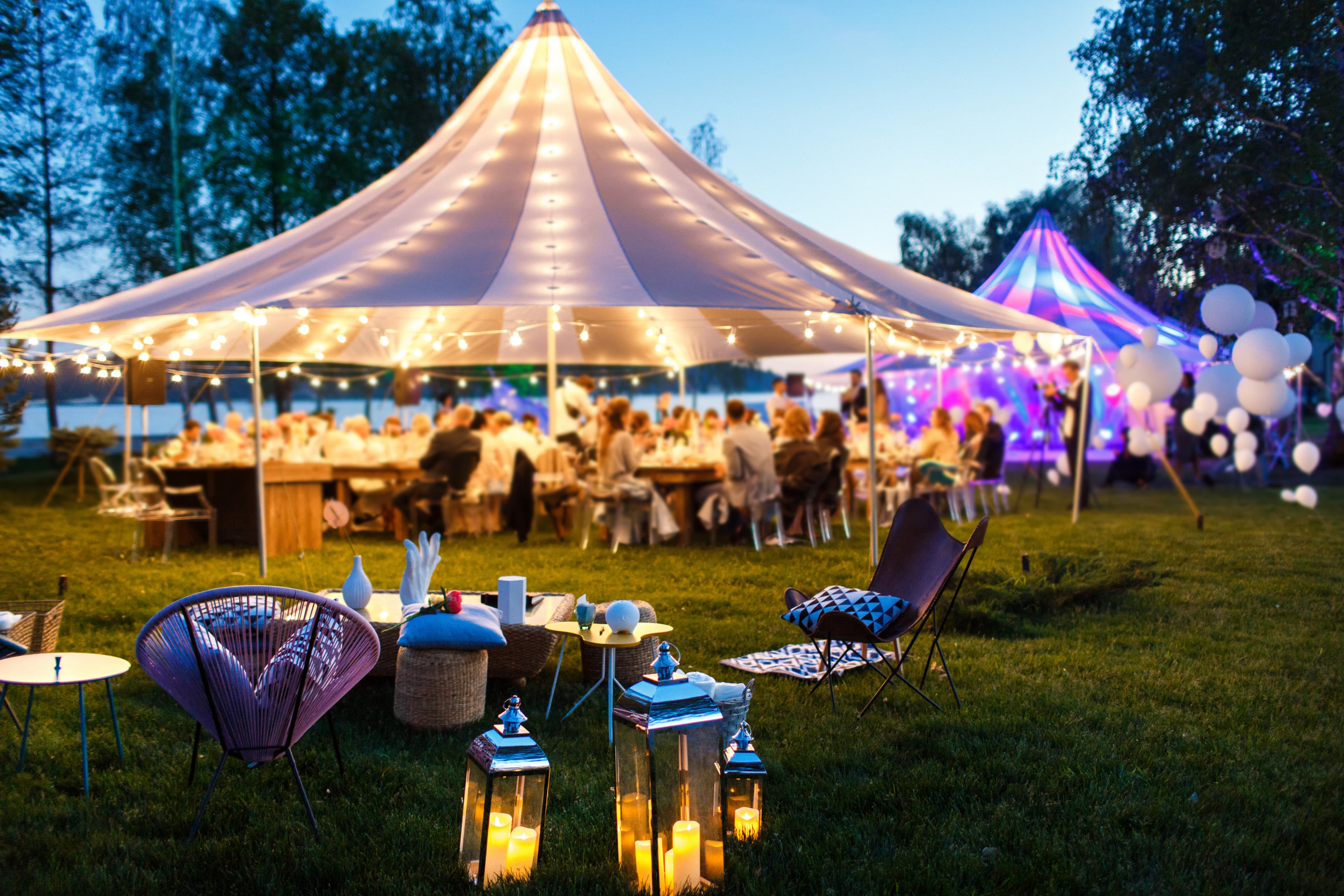 Festively lit wedding tent with guests at dusk on a lakeside meadow.