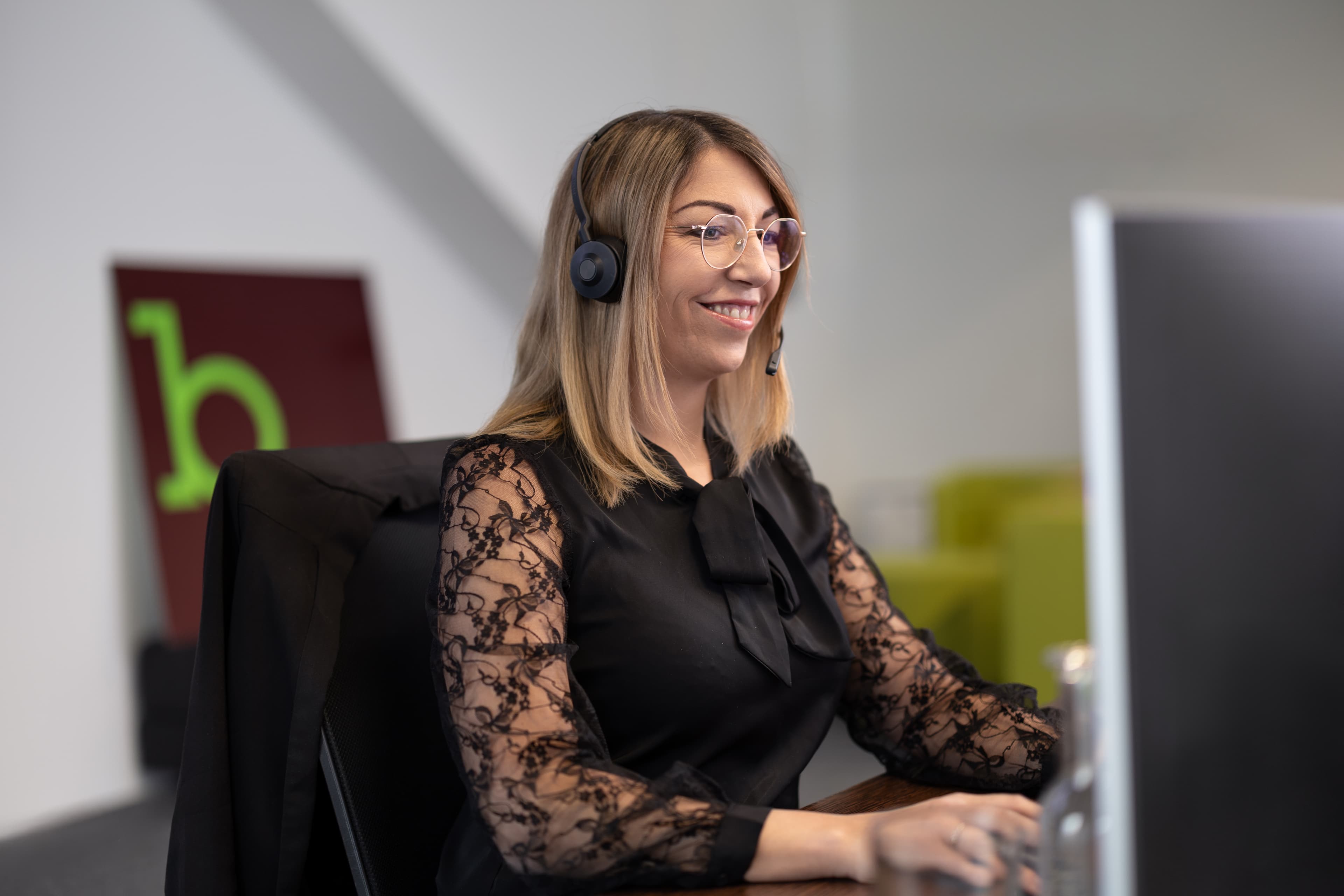 Woman with headset and glasses sits smiling in front of a computer screen.