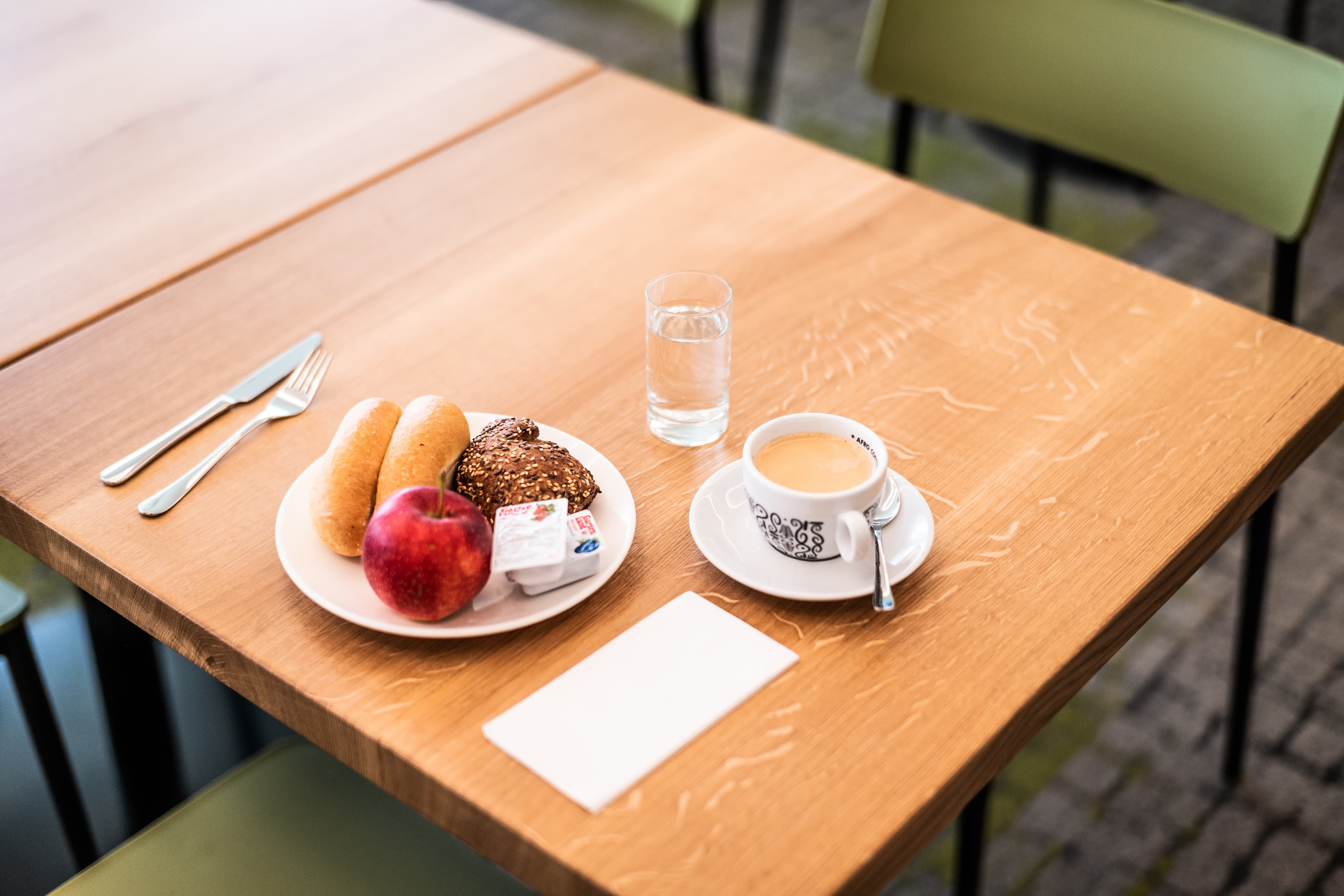 On a wooden table is a breakfast with two rolls, a multigrain roll, an apple, jam, a glass of water, and a cup of coffee at the Flex Motel Spielberg.