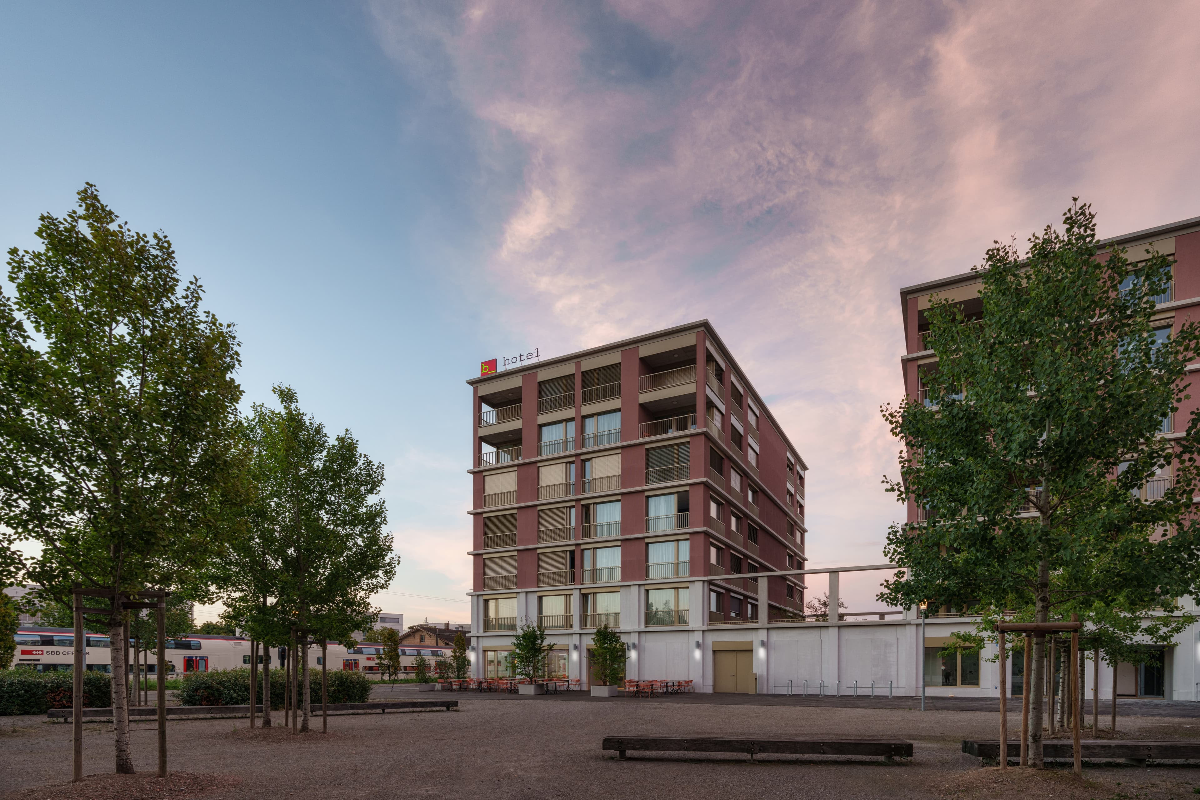 Multistory hotel building with modern balconies at sunset next to trees and a train.