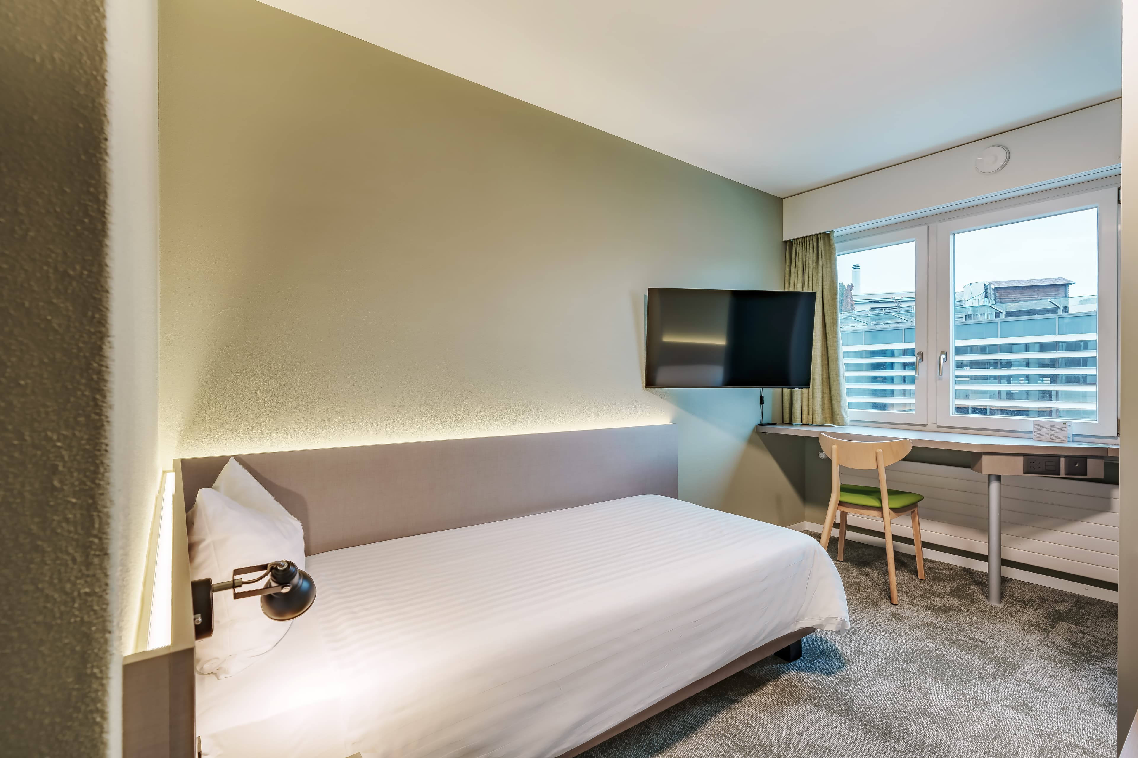 A modern single room at the Flex Motel Wil with a bed, wall lamp, flat screen TV, and desk in front of a window with a view.