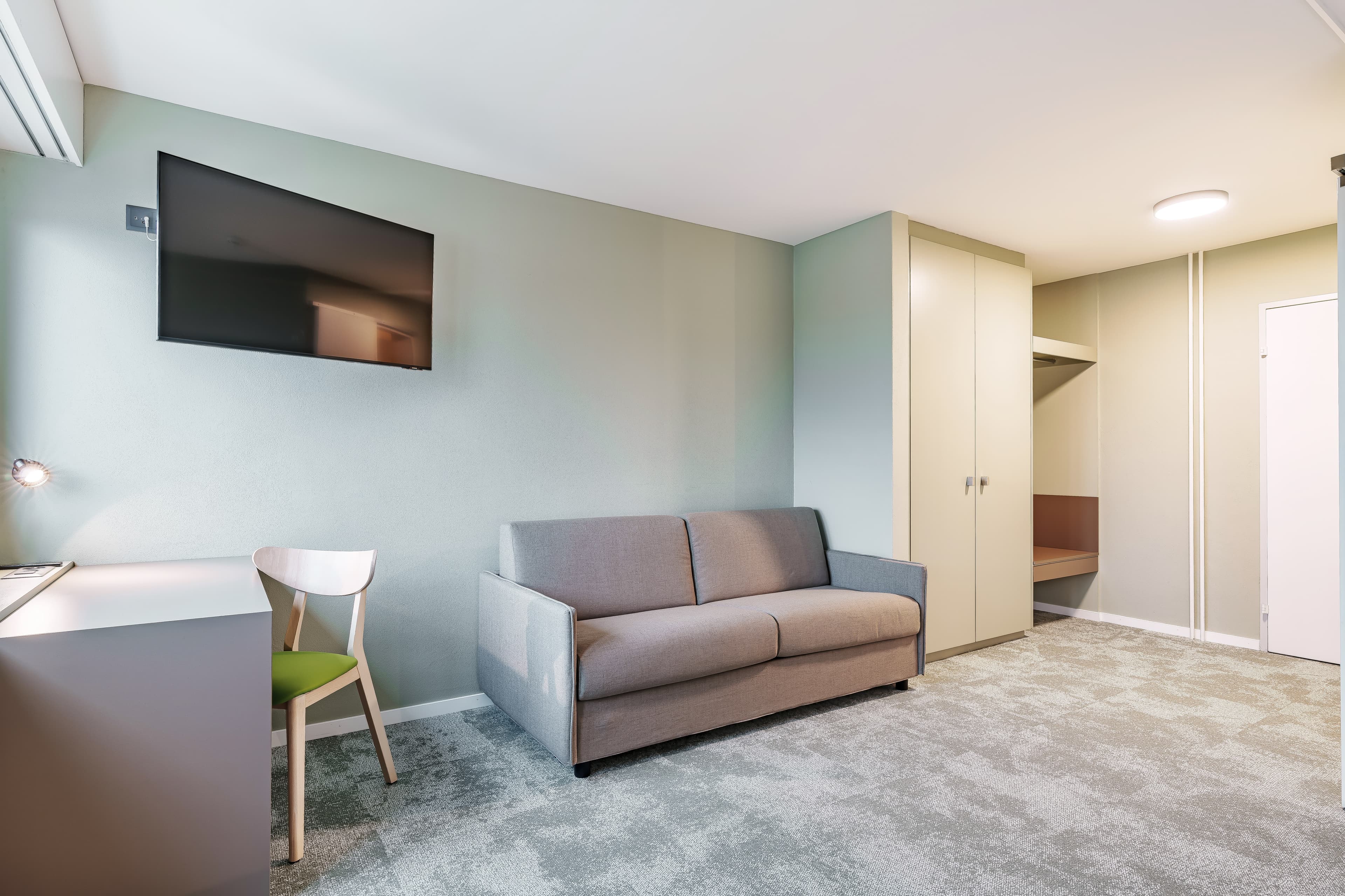 A junior suite at the Flex Motel Wil with a gray sofa, a desk with a chair, a wall-mounted TV, and a wardrobe.