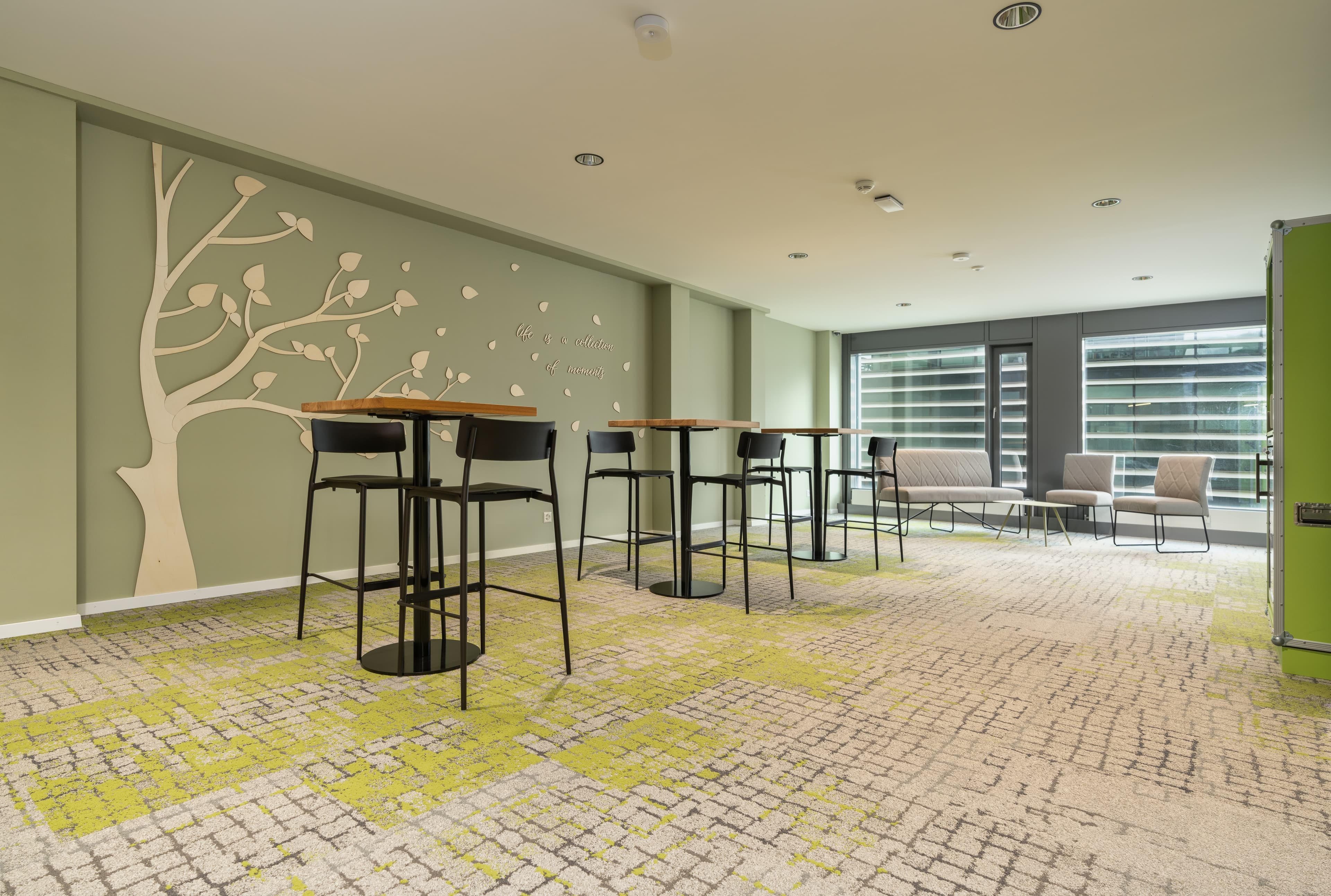 A modern room in the Flex Motel Wil with high tables and chairs, light carpet with a green pattern, and a wall decoration in the shape of a tree.