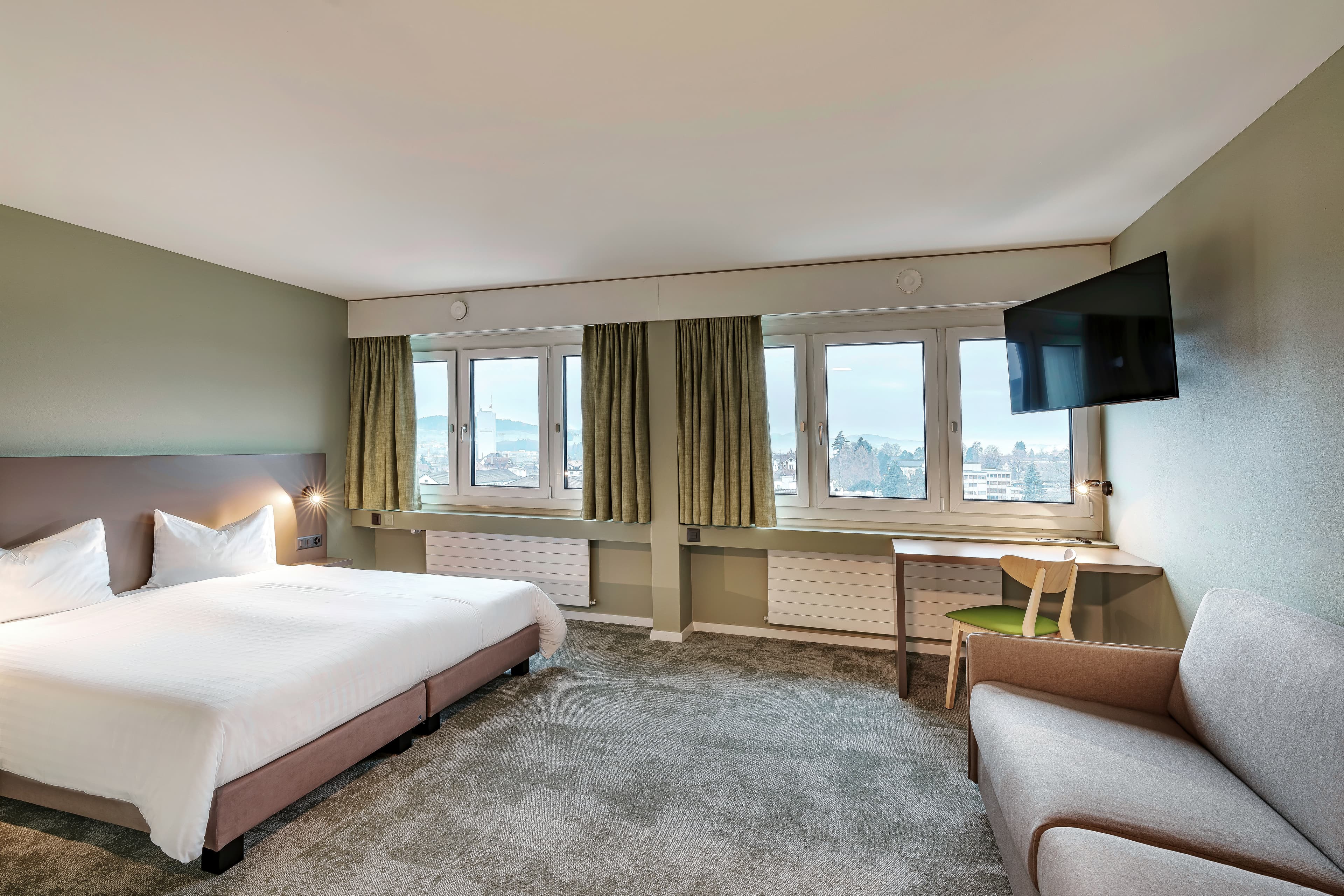 A hotel room at the Flex Motel Wil with a double bed, gray couch, desk, TV, and large windows overlooking the city.