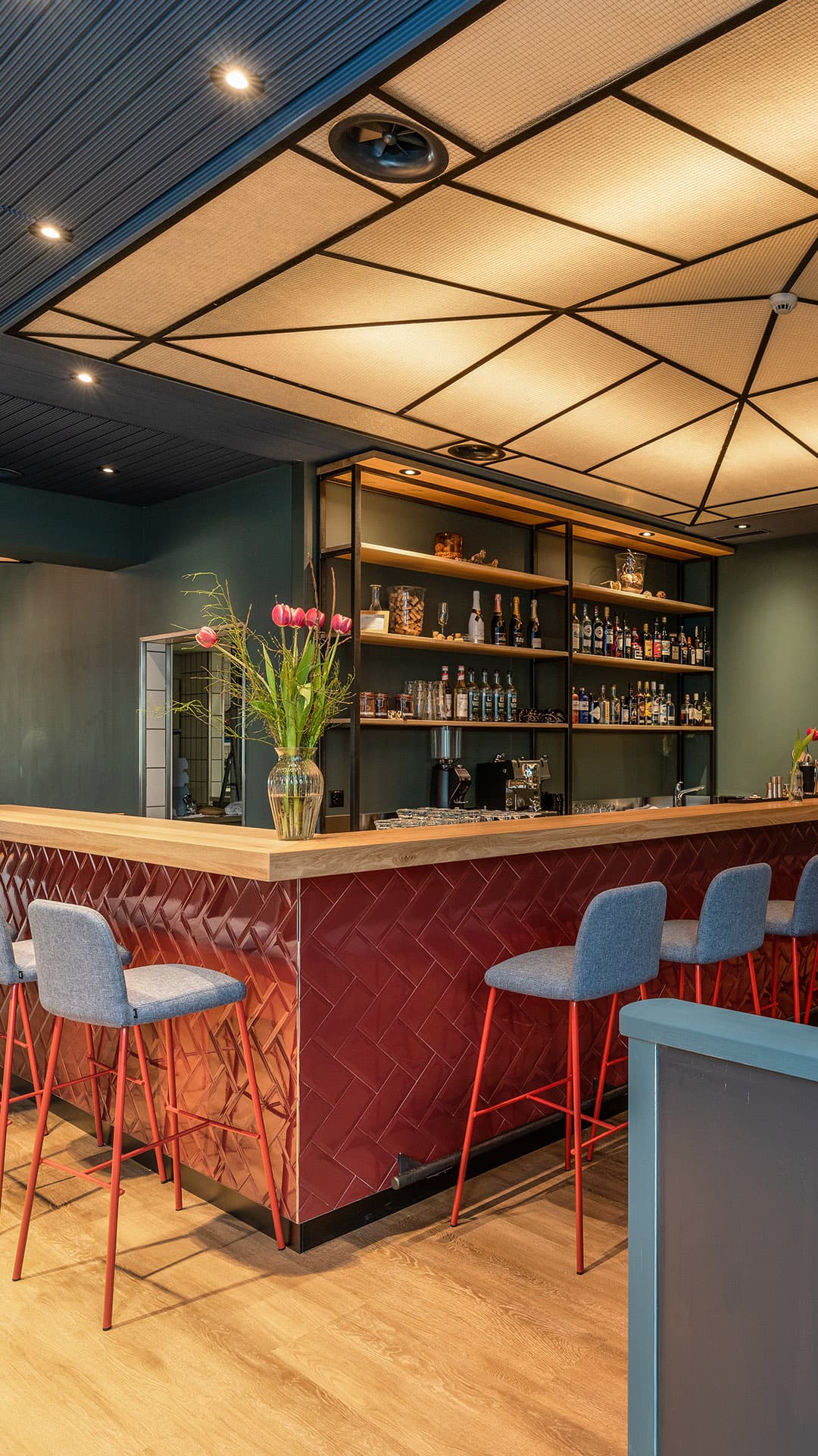 A modern bar in the Schonenwerd restaurant with red tiles gray bar stools and a lit shelf full of bottles