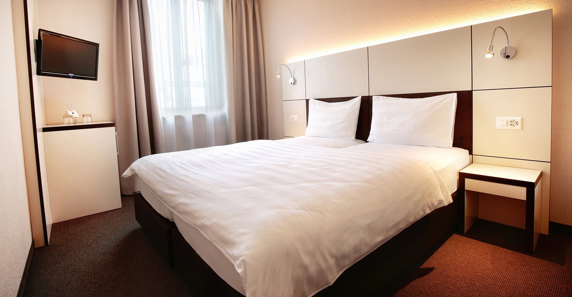 A modern hotel room at the B_Smart Motel Basel with a large double bed, a flat screen TV on the wall, and a stylish nightstand.