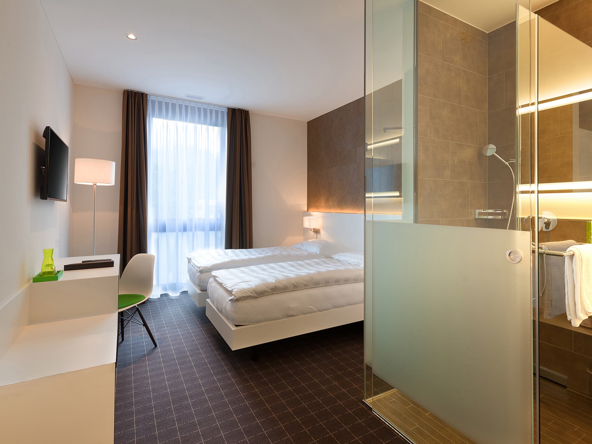 Bright double room in the b smart motel Sevelen with two single beds, large window, desk, chair, television, and an open shower