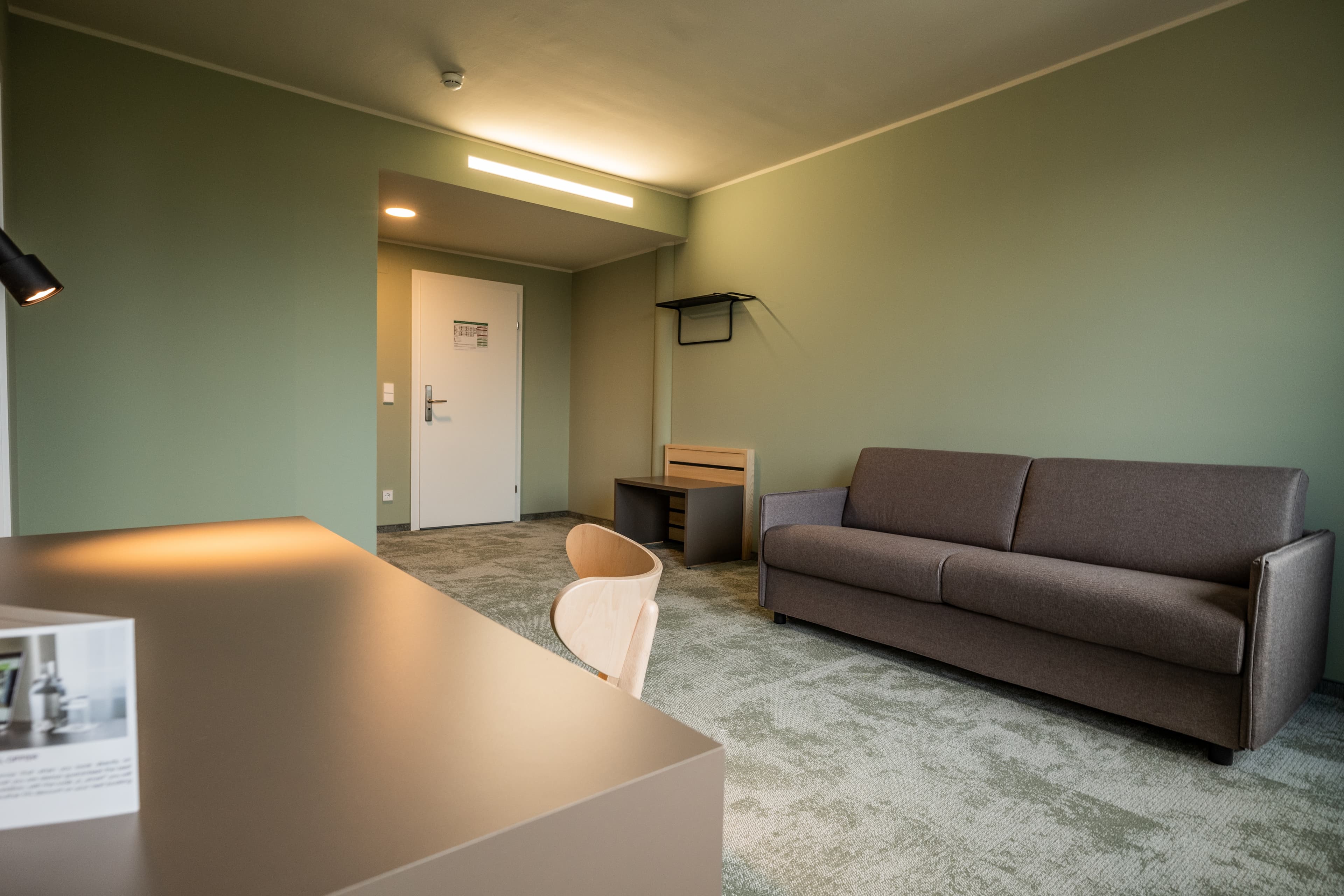 A modern hotel room at the Flex Motel Spielberg with green wall color, a gray sofa, wooden furniture, and a dining table that makes the space inviting and functional.