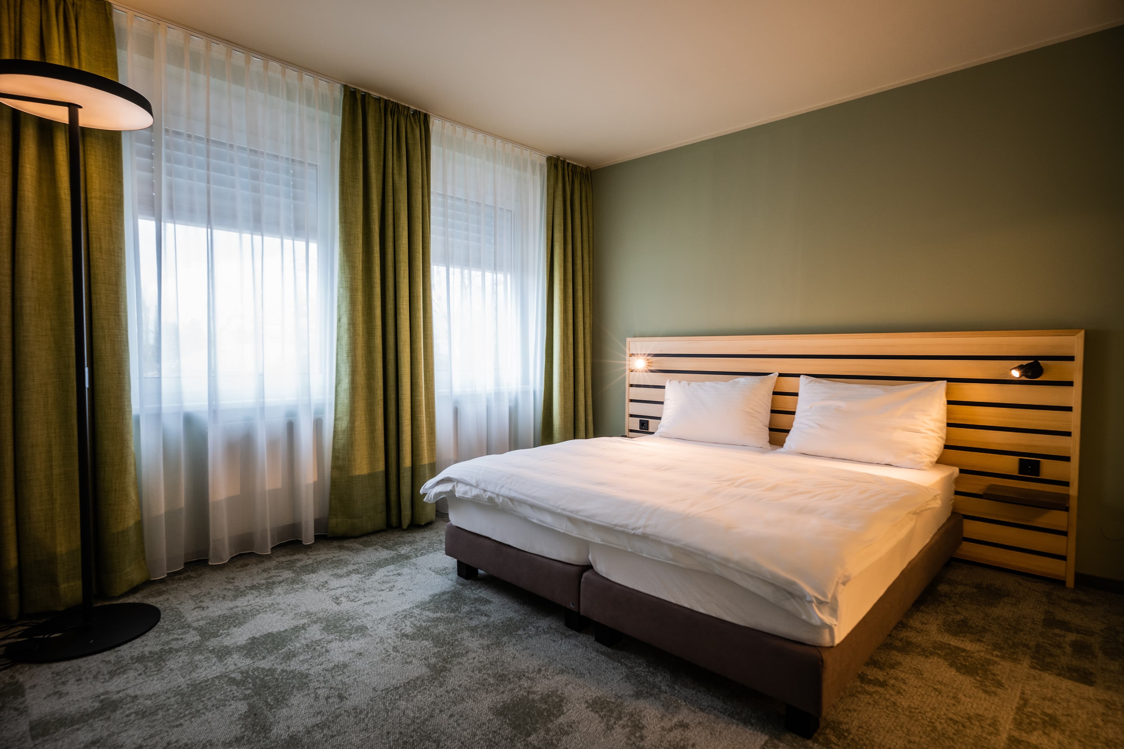 A hotel room of the Junior Suite Standard at the Flex Motel Spielberg with a double bed greenish curtains and a modern floor lamp