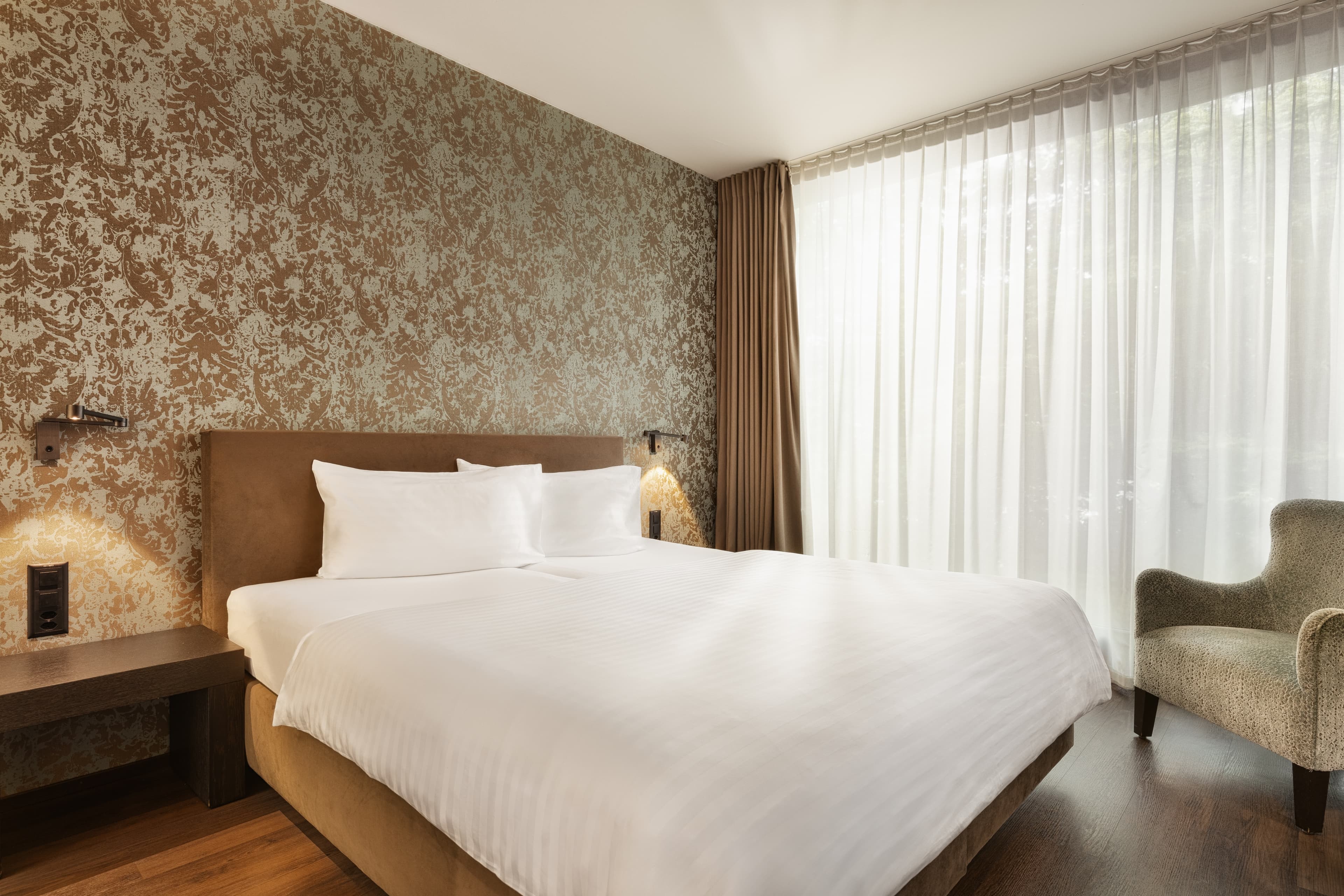 A standard double room at the b_smart Hotel Widnau with a large bed, elegant wallpaper, and a chair next to a large window with white curtains.