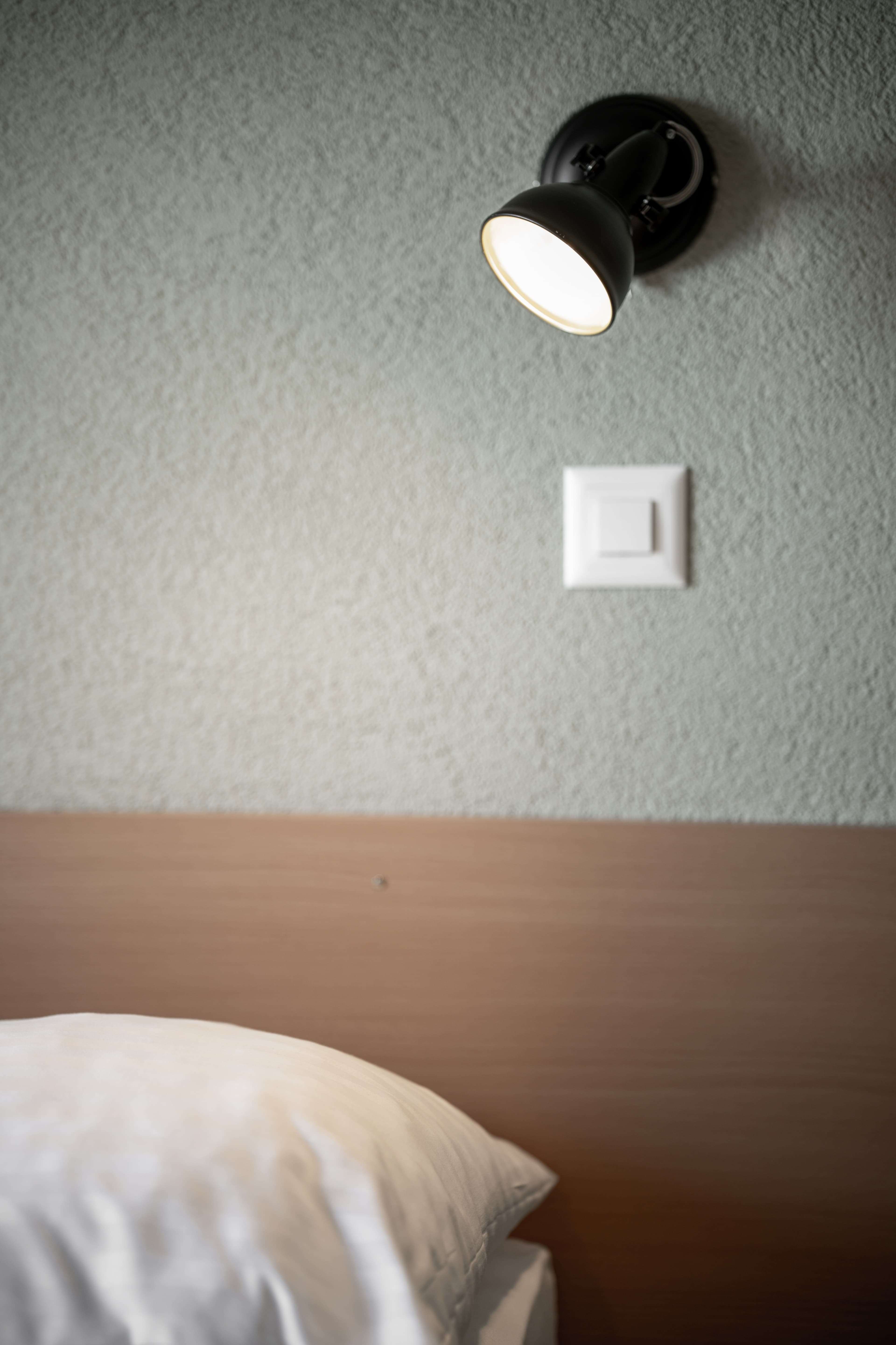 A black wall lamp shines over a white pillow and a wooden bed headboard in the Flex Motel Schonenwerd