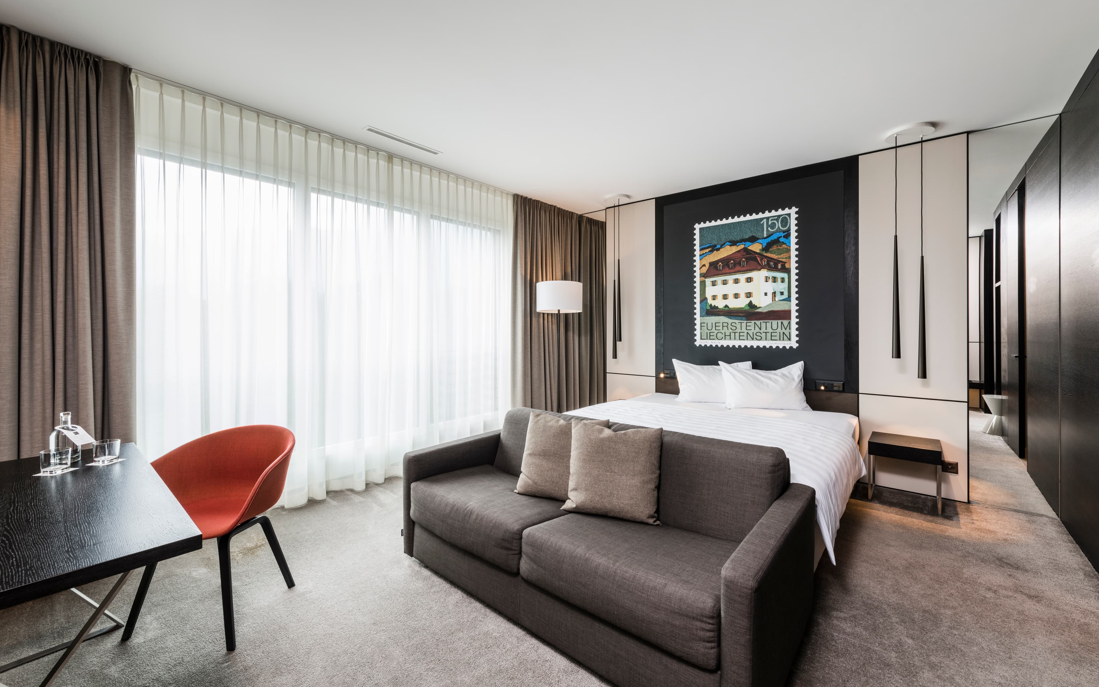 A modern hotel room of the Junior Suite at the b_smart Hotel Bendern equipped with a bed a sofa a desk and a large mural of a postage stamp