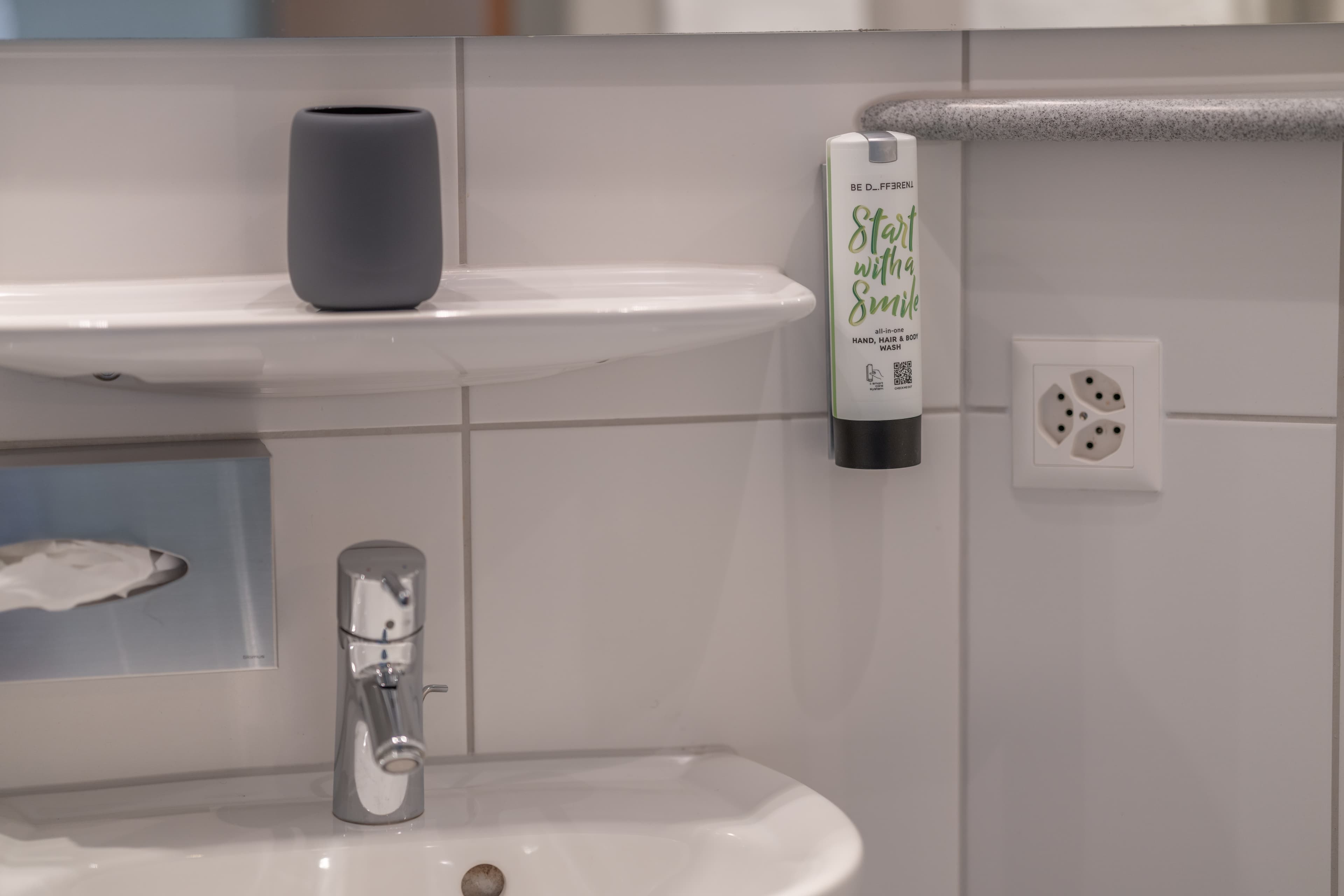 Sink area of a bathroom with a soap dispenser labeled Start with a Smile a cup and an electrical outlet