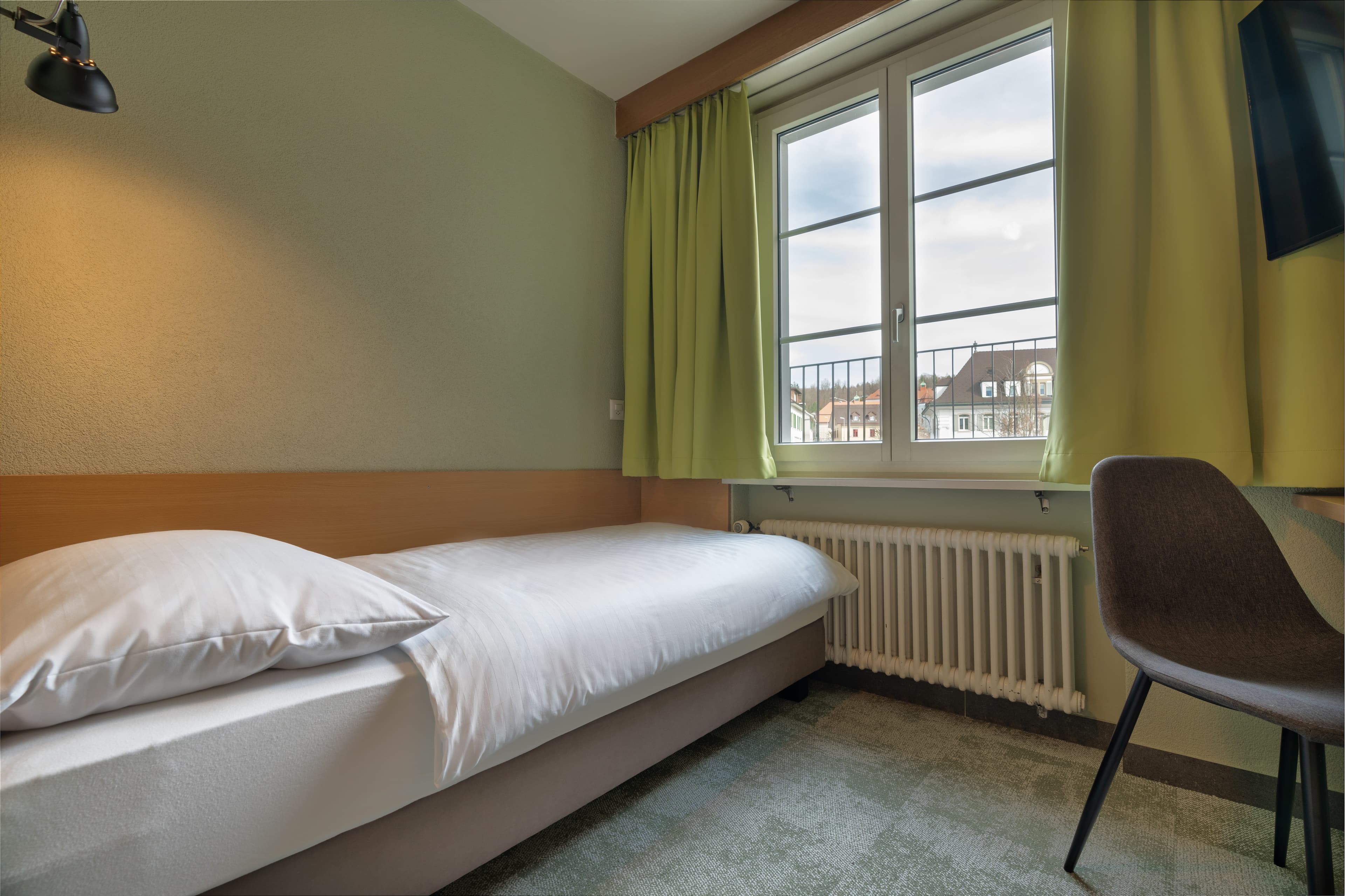 A single room in the Flex Motel Schonenwerd with a single bed green curtain window with view of houses and a chair